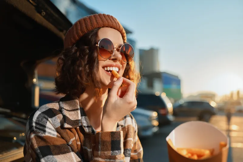 Woman eating fries by car trunk at sunset with sunglasses and smile, candid lifestyle portrait showing authenticity and golden hour glow, mindful living and emotional storytelling vibe.