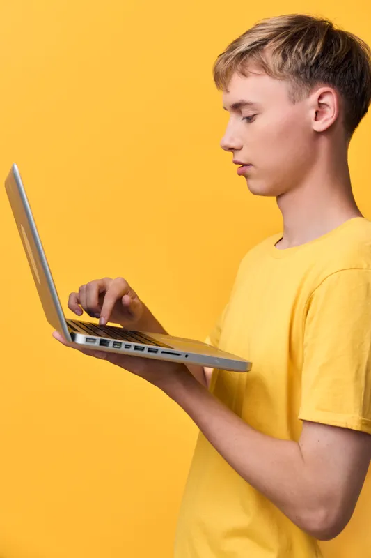 Boy in a yellow shirt typing on a laptop against a bright yellow background, showcasing technology use, learning and concentration in a modern study moment