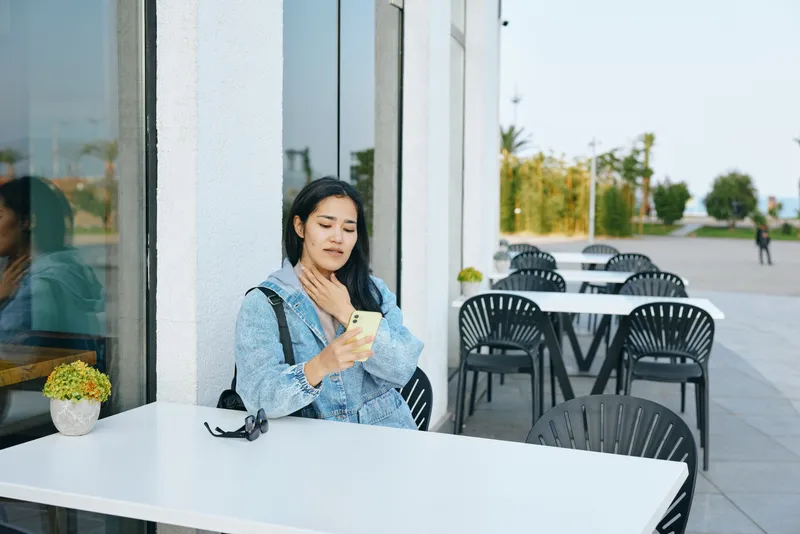 Woman with denim jacket using cellphone at outdoor cafe, sunglasses on table, glass storefront, calm moment of casual browsing and relaxed work in a modern patio setting