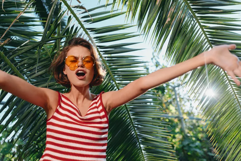 A cheerful traveler enjoys sunny outdoors with arms spread wide, wearing a red striped tank top and orange sunglasses among palm trees, conveying energy and carefree vibes.