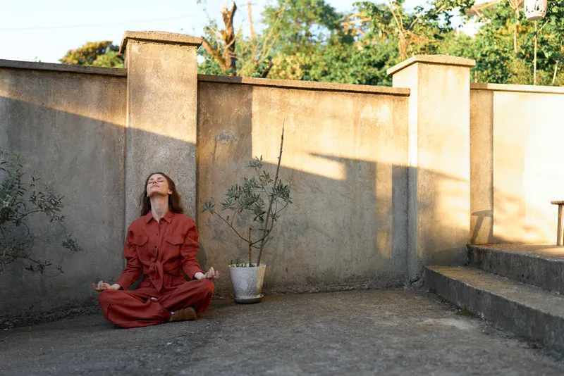 A calm woman in a red jumpsuit sits cross-legged against a sunlit concrete wall, meditating in a quiet courtyard, as soft shadows from nearby planters fall around her