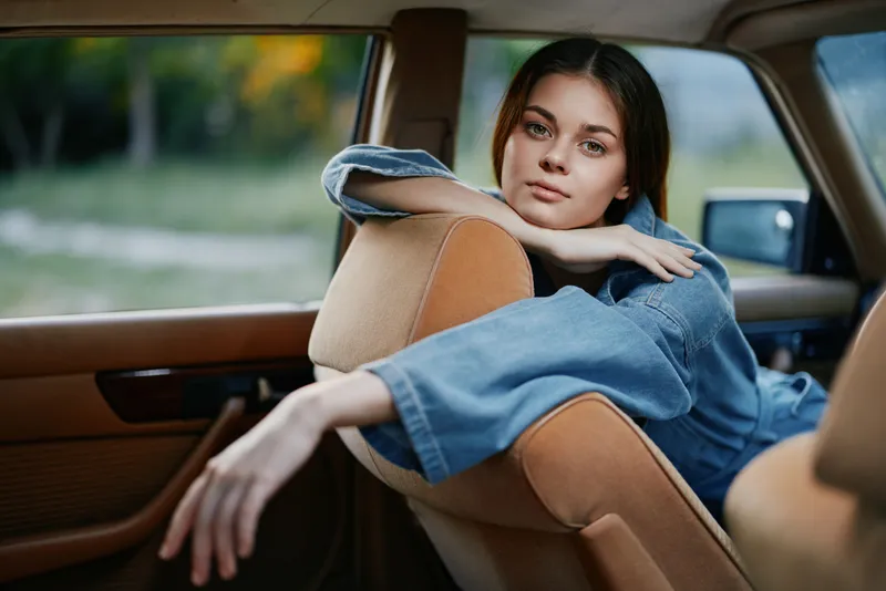 Young woman in a vintage car wearing denim, relaxed pose, smiling softly, green and golden nature background, capturing a sense of nostalgia and freedom.