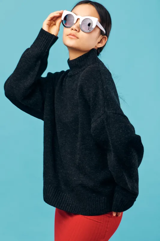 Woman wearing oversized black sweater and red skirt posing with white sunglasses on blue background. Fashion portrait with casual stylish outfit and confident pose in studio lighting.