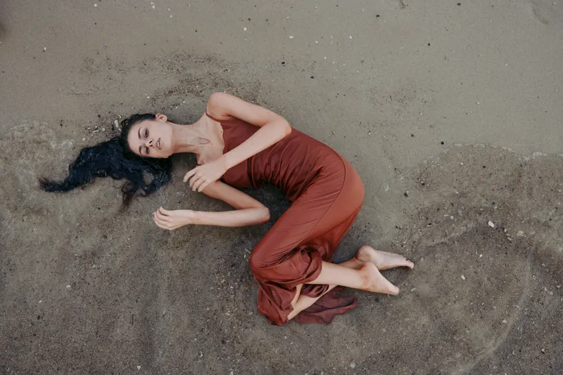Relaxed woman lying on sandy beach with long dark hair, wearing a red dress, expressing calm and serenity in a natural setting.