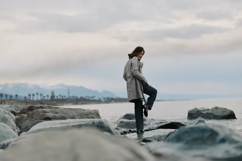 Woman in grey coat stands on rocky shore by the sea, balancing on one leg. Overcast sky and distant mountains create a calm, contemplative outdoor atmosphere.