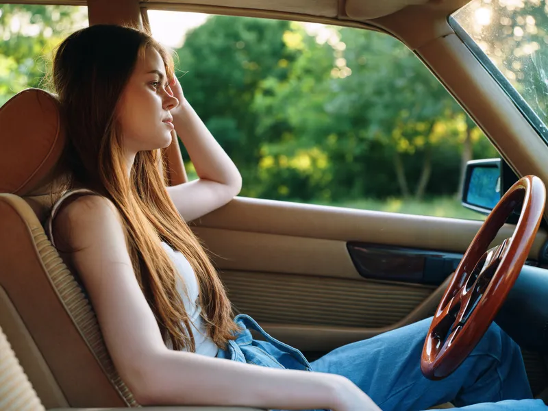 Young woman with long hair sitting in a car, feeling thoughtful against a natural green background, embodying freedom and reflection in casual summer attire.