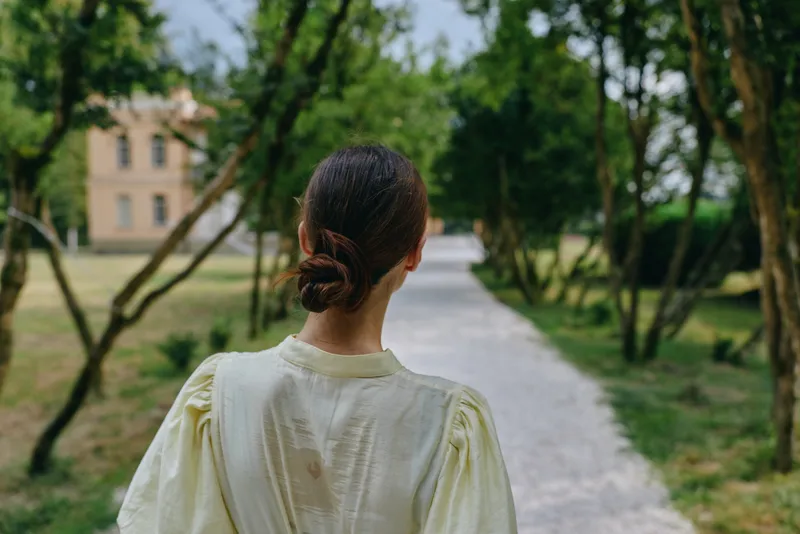 Woman in a soft yellow dress walks away along a tree lined park path, back facing camera, enjoying a peaceful outdoor moment with calm atmosphere and gentle natural light.