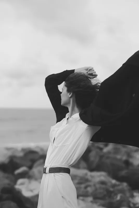 Woman with flowing scarf at seaside, white shirt and belt, arms raised against wind, black and white photography capturing motion and open horizon, dramatic light on rugged rocks