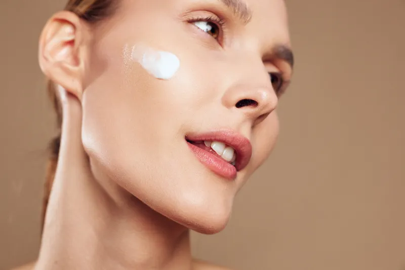 Close up of a young woman applying moisturizing cream on her cheek with smooth skin and natural makeup against beige background. Concept for skincare and beauty routine.