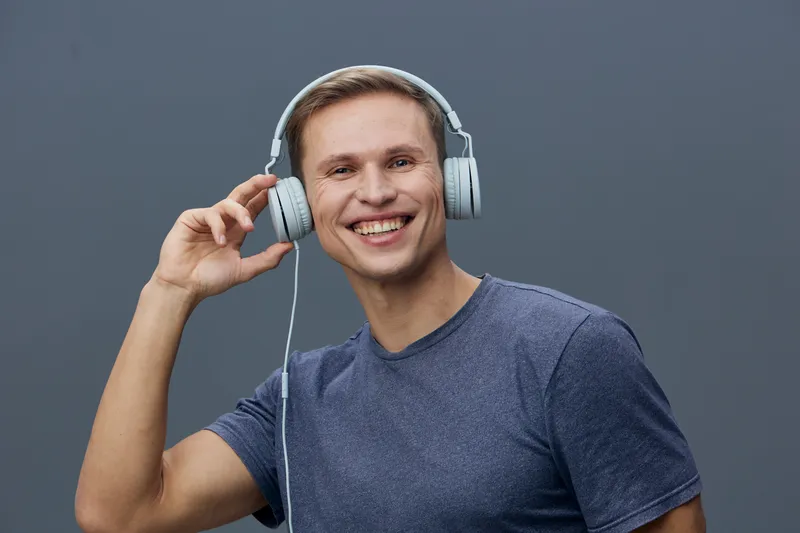 Joyful young man wearing headphones smiling at camera holding music device in hand studio portrait on plain gray background. People lifestyle concept, entertainment, happiness, modern technology, casu