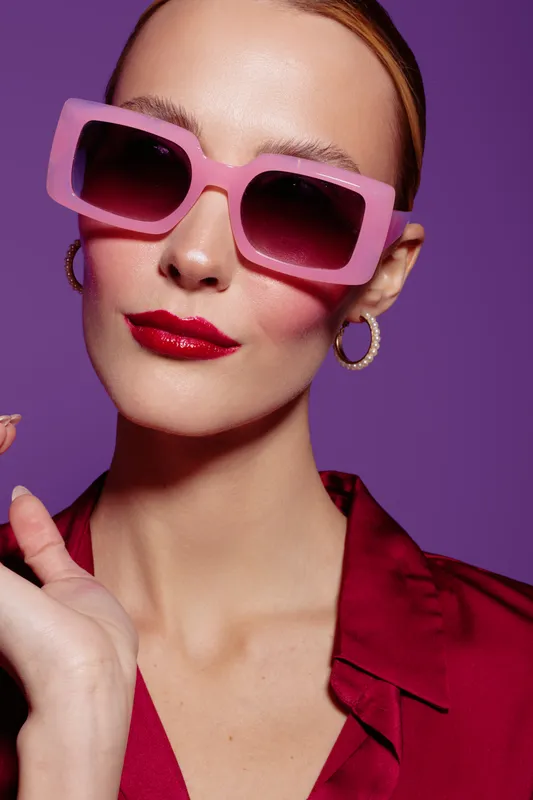 Stylish woman with pink sunglasses and red lipstick poses against purple background. Fashion portrait with trendy accessories and confident expression. Concept of glamour and style.