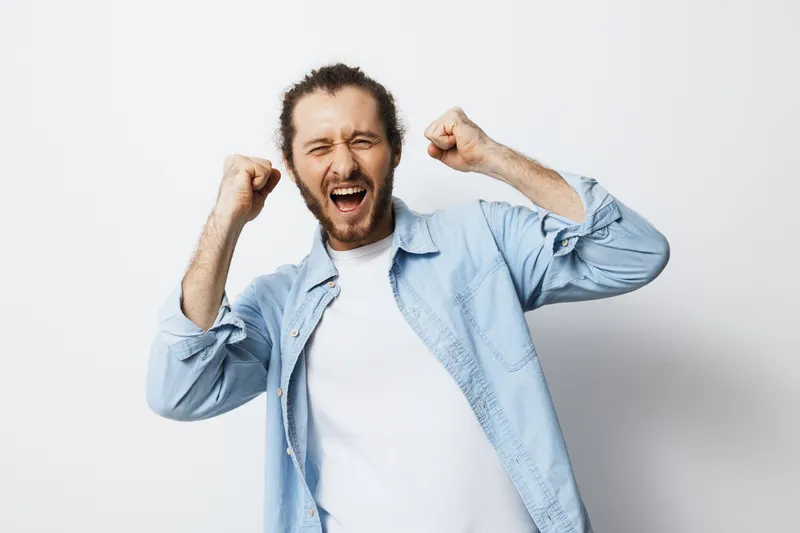 Happy young man celebrating success with raised fists, wearing a light blue shirt against a minimalistic white background, conveying joy and achievement