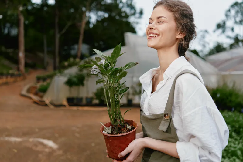 Happy young woman holding a potted plant in a greenhouse, showcasing her joy for gardening and nature, dressed in casual attire