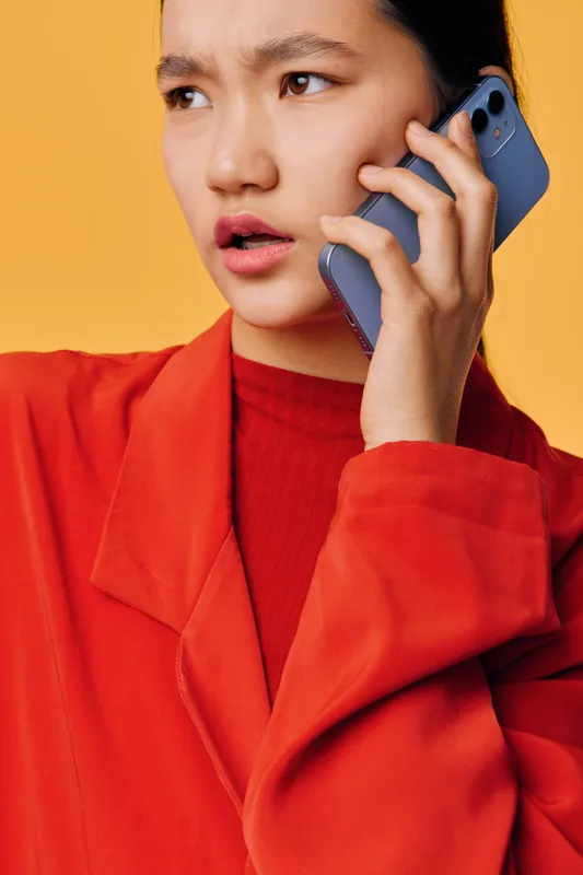 Young woman in red jacket speaking with smartphone on ear with serious face expression against yellow background. Closeup portrait of girl using mobile phone for communication.