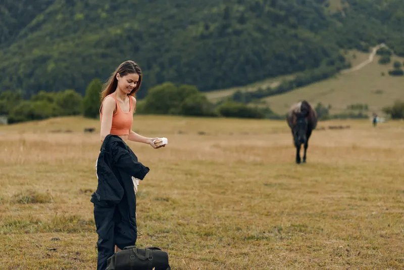 A young woman stands in a wide grassy field with a horse in the distance, adjusting her gear and smiling, capturing a moment of outdoor peace, exploration, and calm rural atmosphere.