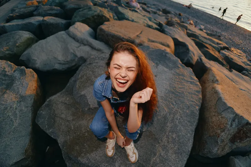 Lifestyle woman in modern boho-western denim street style laughing joyfully on rocky beach during golden hour with film color tone