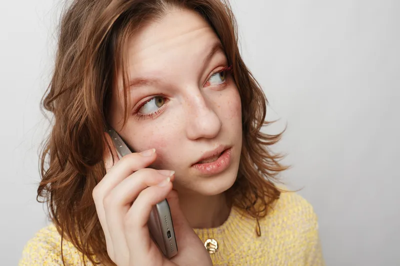 Young woman with freckles and brown hair talks on smartphone looking aside. Casual style, yellow sweater, natural light. Concept of communication, technology, and thoughtful expression.