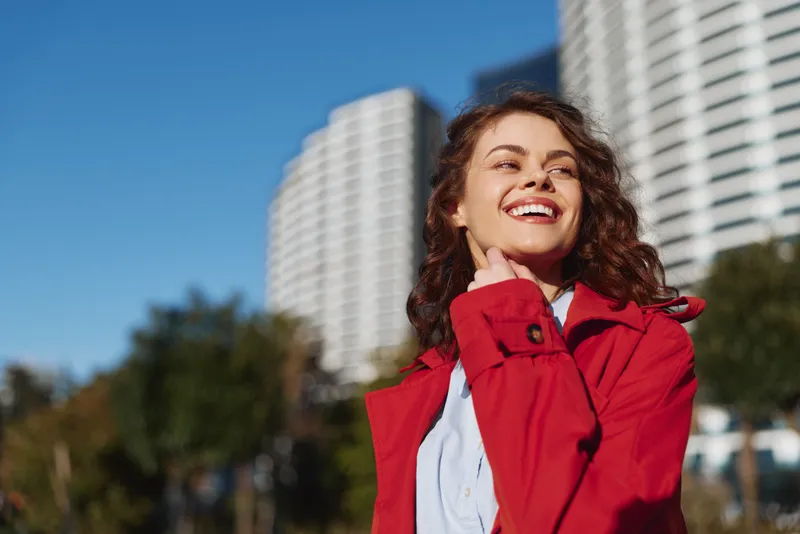Smiling young woman with curly hair in a red jacket poses outdoors in an urban setting, with tall buildings and a bright blue sky behind.