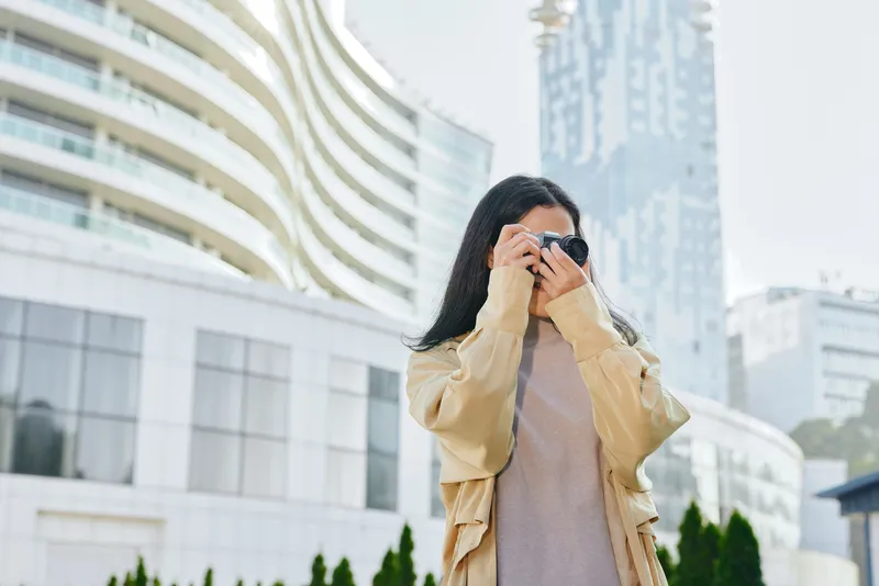 Woman adjusting sunglasses while holding a camera in an urban outdoors scene with modern architecture, showcasing street fashion, coat style, and city lifestyle amid daylight ambience