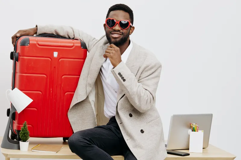 Smiling man with dark skin wearing sunglasses and a light grey blazer sitting at a desk with a red suitcase and laptop, happy and confident expression, travel concept, modern office background.