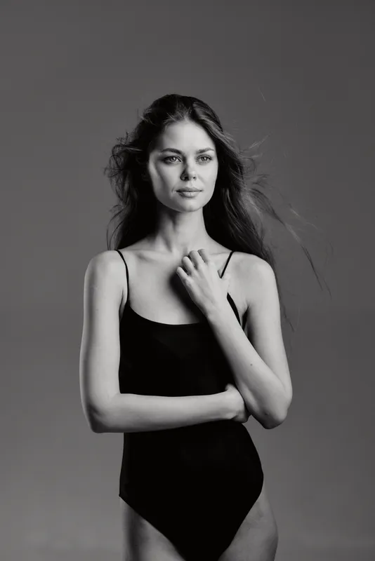 Black and white portrait of a young woman in a simple black bodysuit with calm expression and windblown hair.