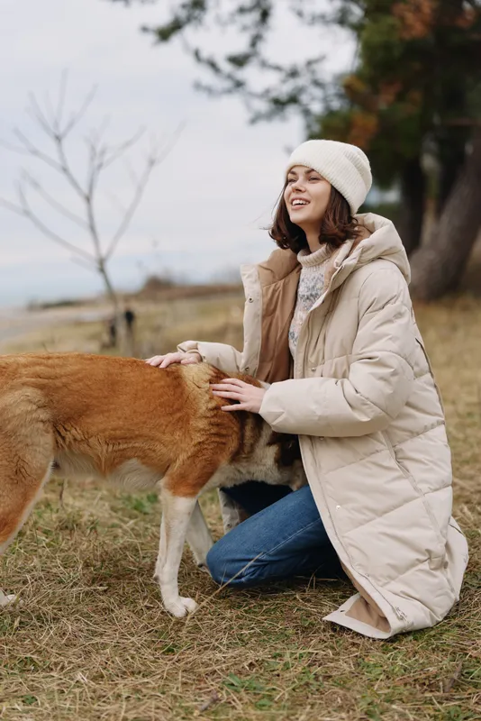 Woman kneeling outdoors in a warm coat and beanie gently pets a dog, amid a grassy field with trees, capturing a candid, joyful moment in nature.
