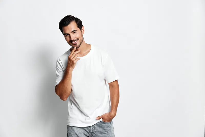Handsome smiling man in casual white t-shirt stands confidently with hand on chin, looking at the camera, isolated on a plain light background. Lifestyle concept.
