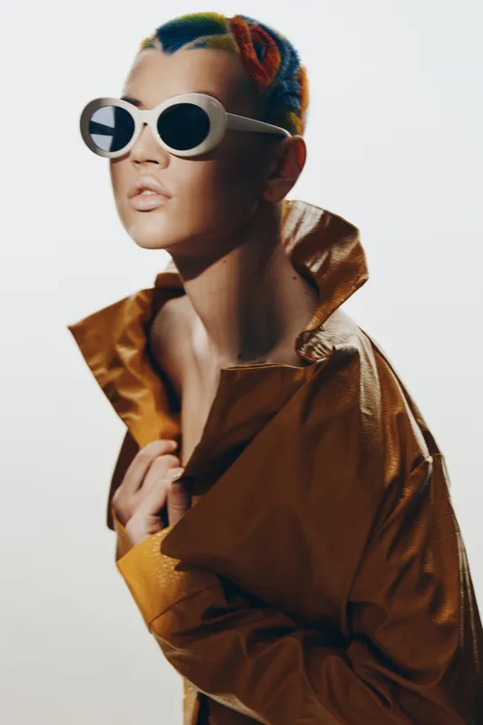 fashion woman with colorful braided hair wearing white sunglasses and a mustard yellow leather jacket, posing confidently against a plain white background