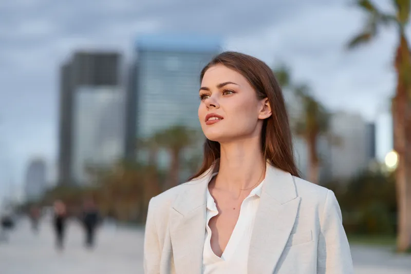 confident business woman in stylish white blazer walking in urban cityscape during daytime with modern buildings and palm trees in background