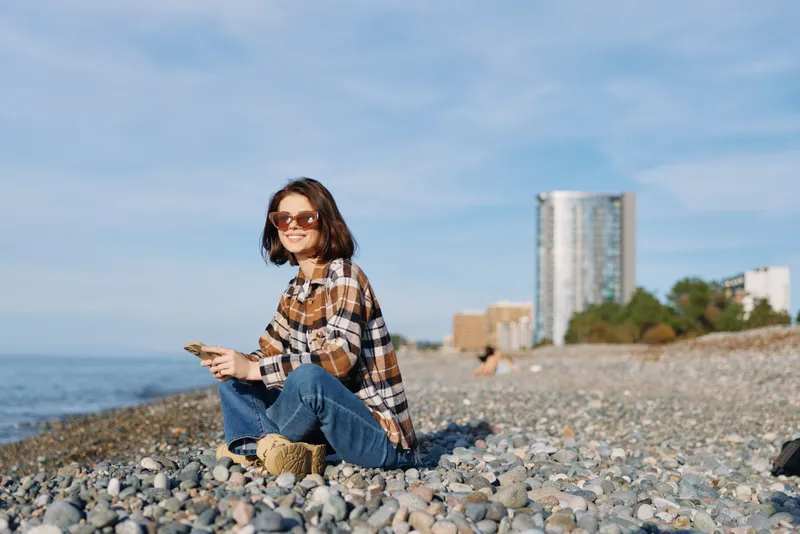 Woman at a pebble beach wearing a plaid shirt and jeans, sitting with sunglasses, enjoying the seaside with a calm smile and distant cityscape.