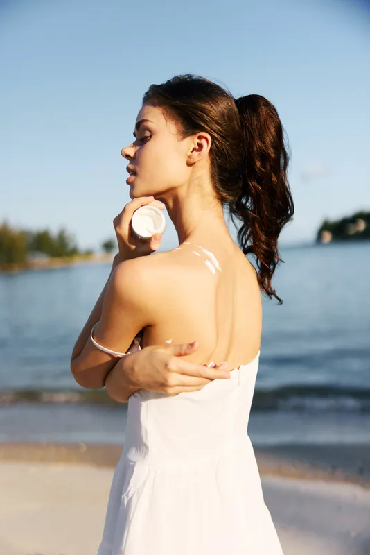 Portrait of a young woman standing on a sunlit shore, turned profile, wearing a white dress, holding a seashell near her shoulder as gentle waves shimmer.