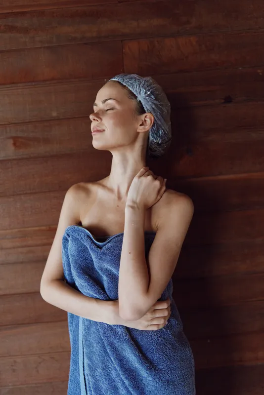 Relaxed woman wrapped in blue towel with shower cap, enjoying calm moment against wooden wall in spa-like environment, soft natural light highlighting serene mood.