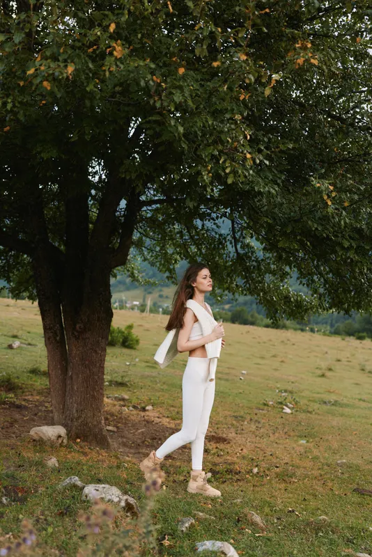 Woman in white outfit walking beside a large tree in an open field, capturing relaxed outdoor lifestyle, nature connection, and carefree mood. Soft lighting, warm tones.
