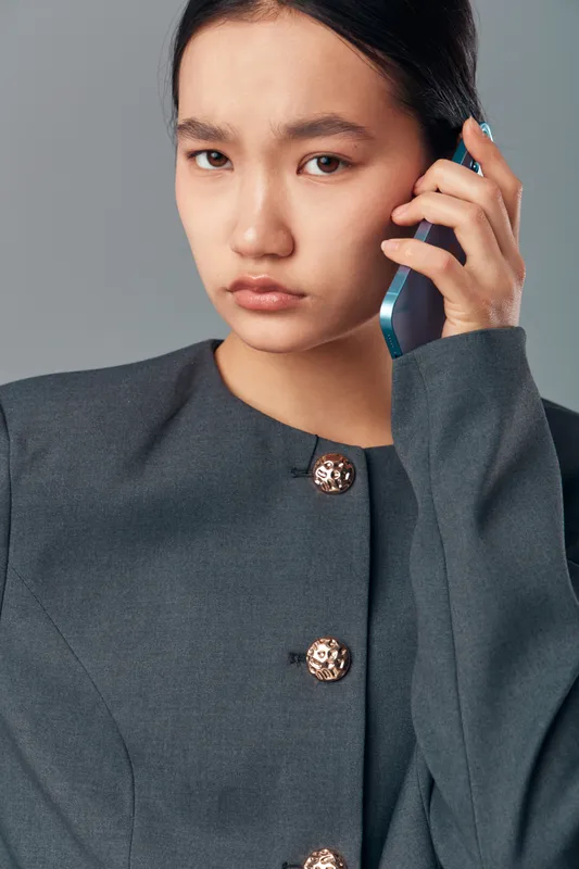 Young woman in gray jacket holds smartphone near ear with serious expression, communication and technology concept for business or personal conversation in studio setting.