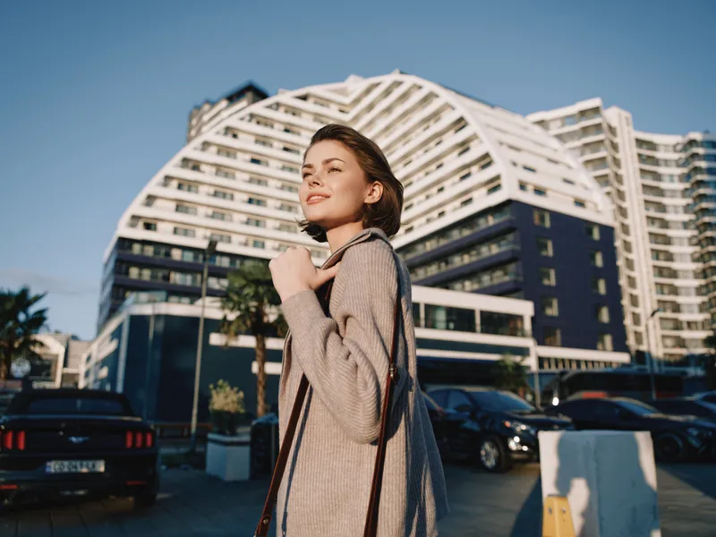 A stylish young woman stands outdoors near a modern urban complex, smiling softly as she gazes ahead, conveying confidence and a relaxed, positive urban lifestyle.