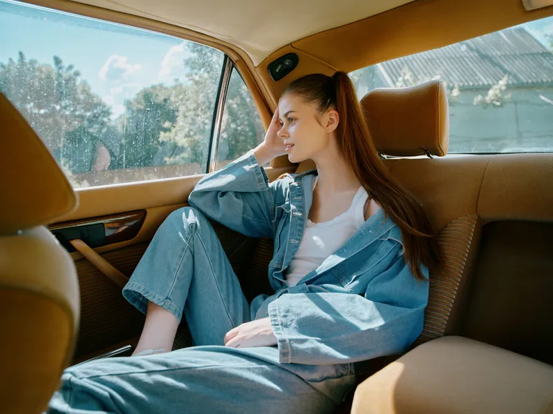 Thoughtful young woman in casual denim outfit sitting in a car, looking out the window in a serene moment, reflecting on her thoughts amidst a soft sunlight.