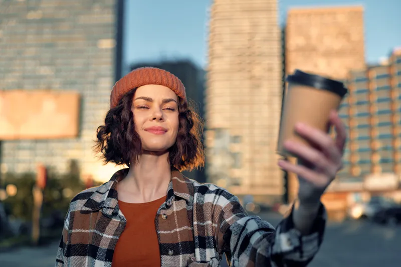 Woman with coffee in city at golden hour, smiling with eyes closed, beanie and plaid shirt, candid moment of authenticity and mindful living, emotional storytelling with warm golden glow.