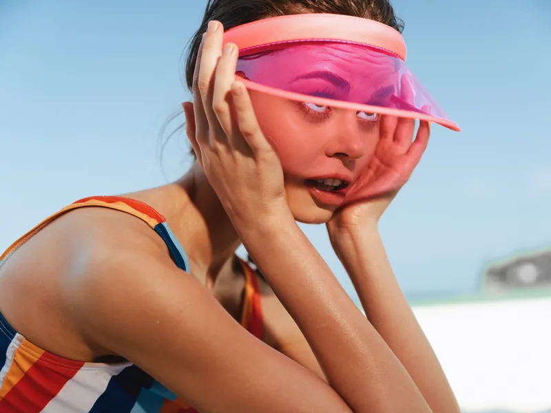 A smiling woman wearing a bright striped swimsuit and a pink visor at the beach, adjusting hair as sunlight highlights sunlit skin and a playful summer vibe.