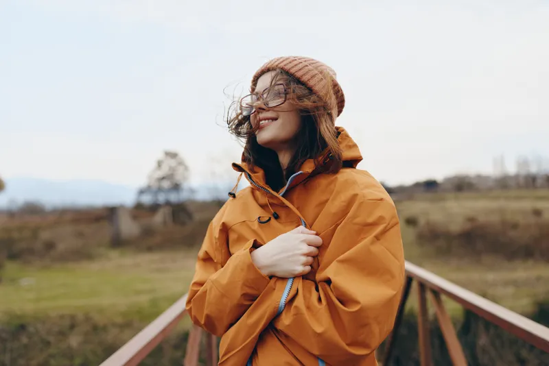 Woman lifestyle portrait outdoors on rural walkway, wearing orange jacket, knitted hat and scarf, smiling and looking at distance, windy day, holding hands near coat, soft natural light