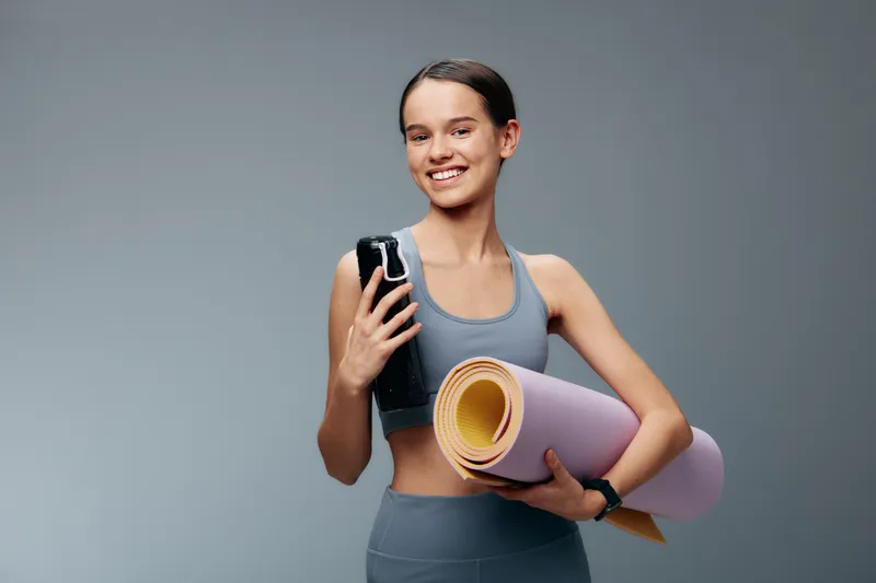 Young woman in athletic wear holds a water bottle and rolled yoga mat, ready for a workout in a simple studio. Emphasizes fitness lifestyle, training and balance for a healthy routine.
