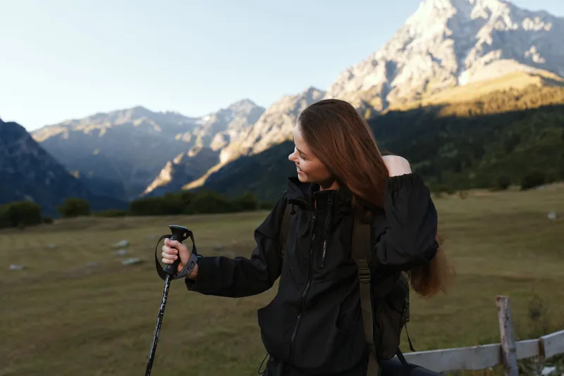 A young hiker in a black jacket holds a trekking pole in a scenic valley with towering peaks. She smiles softly, enjoying the outdoors and a calm moment in nature.