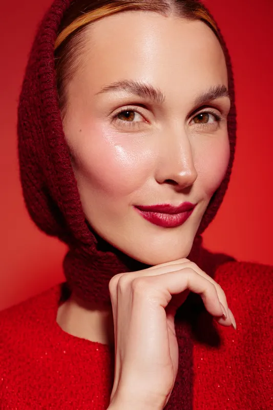 Close-up portrait of a confident woman wearing a red knitted hood and sweater, with glossy skin and red lipstick, posing against a red background. Concept of fashion and beauty.