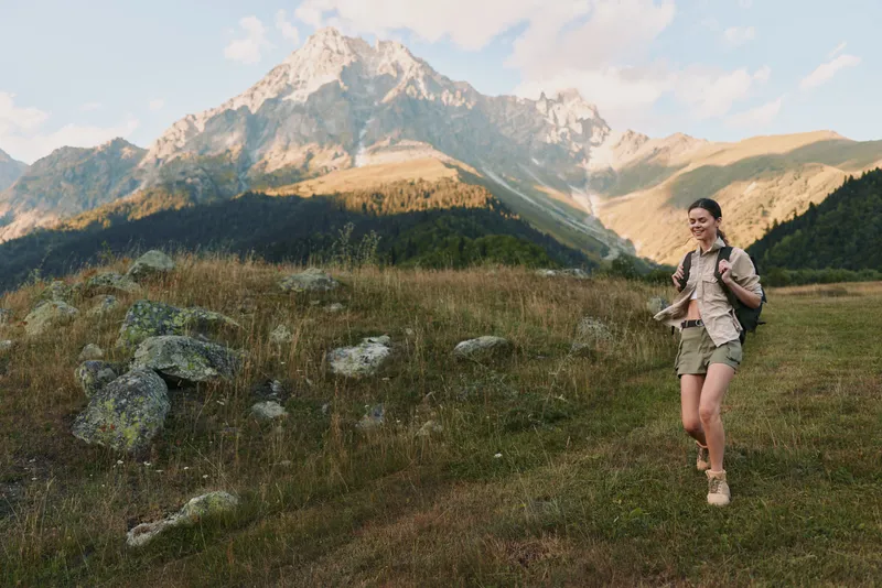 Young woman hiking through a grassy meadow with rugged mountains in the background, smiling and focused on the trail, carrying a backpack and enjoying an outdoor adventure