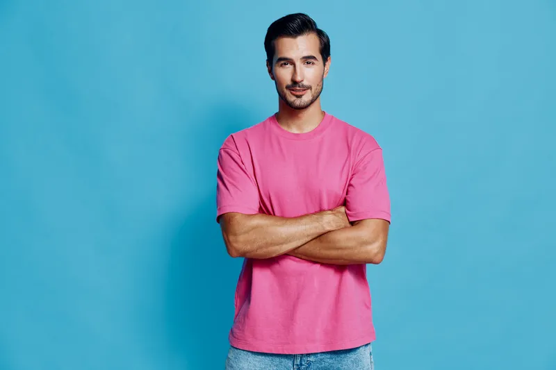Handsome smiling man wearing a pink t-shirt, standing confidently with crossed arms against a bold blue background. Modern lifestyle concept.