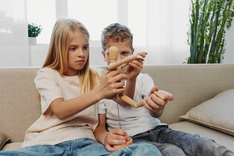 Two focused children playing with wooden mannequin on couch in bright living room, exploring creativity and imagination through hands-on activity.