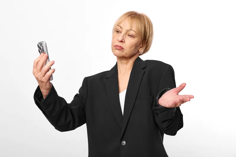 Confused woman in business blazer holding smartphone in one hand, making a gesture with open palms, studio portrait on white background, neutral tones