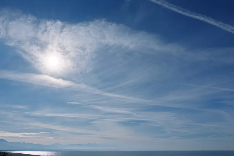 Clear blue sky over calm sea with bright sun and wispy clouds, expansive horizon and reflective water surface creating serene coastal atmosphere, ideal for nature and travel themes