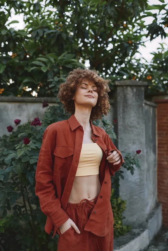 Curly haired woman in a rust suit and beige crop top stands outdoors with eyes closed enjoying warm sunlight