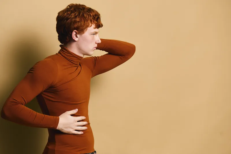 Young man with red hair in brown turtleneck posing sideways against beige background with hand on stomach and other behind head in thoughtful posture.