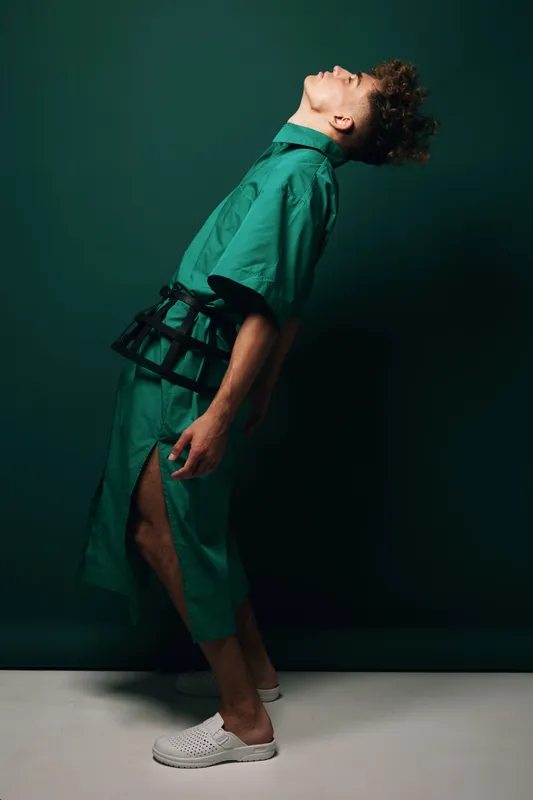 Man with curly hair wearing oversized green pareo skirt with large slit and green shirt with wide sleeves bending backwards against plain studio green background.